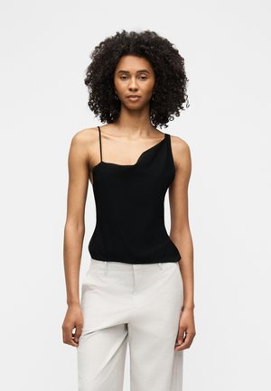 Femme aux cheveux bouclés portant un haut sans manches noir asymétrique et un pantalon taille haute beige clair, debout devant un fond uni.
