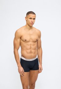 Μυώδης νεαρός με κοντά μαλλιά που φοράει μαύρα εσώρουχα Tommy Hilfiger, στέκεται μπροστά σε απλό φωτεινό φόντο.