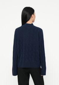 Persona con cabello largo y negro, vistiendo un suéter de punto azul marino y pantalones negros, de pie de espaldas contra un fondo blanco liso.