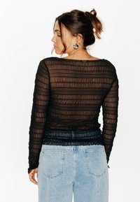 Top nero trasparente a maniche lunghe con texture arricciata, scollo arrotondato e vita aderente. Abbinato a jeans azzurri a vita alta.