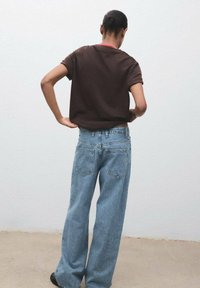 Camisa marrón de manga corta con cuello rojo, combinada con pantalones vaqueros de pierna ancha en azul claro que presentan detalles texturizados y bolsillos traseros.