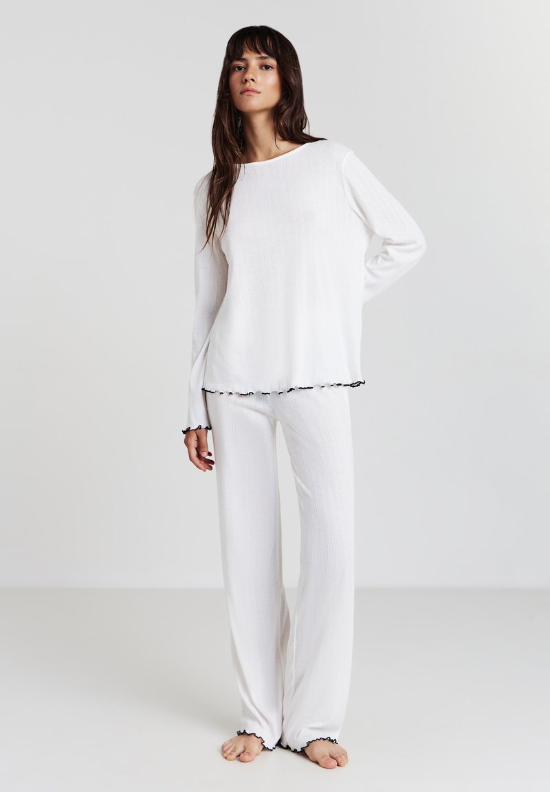 Femme debout pieds nus portant un haut à manches longues blanc côtelé et un pantalon avec une bordure noire sur les poignets et l'ourlet, devant un fond uni.