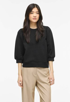 Giovane donna con capelli lunghi e scuri, indossa una maglia nera con maniche a sbuffo e pantaloni beige, in piedi davanti a uno sfondo bianco semplice.