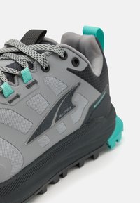 Sive tekaške čevlje za trail s mrežastimi in gumijastimi materiali. Imajo strukturirano zgornji del, črne poudarke in teal podrobnosti na peti in vezalkah.