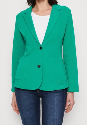 Blazer en coton vert avec une coupe ajustée, revers crantés, deux poches avant et une fermeture à un bouton. Associé à un top blanc et un jean sombre.