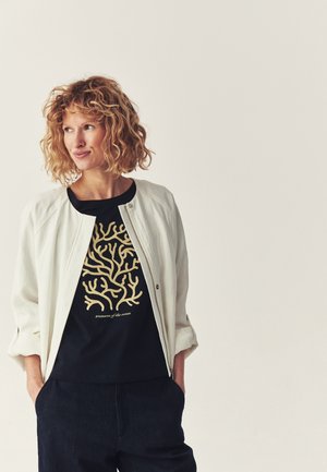 Kobieta o kręconych blond włosach, ubrana w białą kurtkę na czarnej koszulce z złotym wzorem korala i napisem „skarby oceanu.”