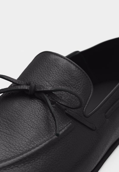 Mocassin en cuir texturé noir avec un détail de nœud sur le dessus, présentant une finition lisse et polie ainsi qu'une conception à bout rond.