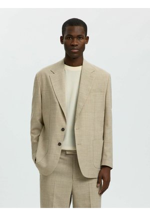 Jeune homme portant un costume beige à deux boutons et un pull blanc texturé, debout devant un fond clair uni.