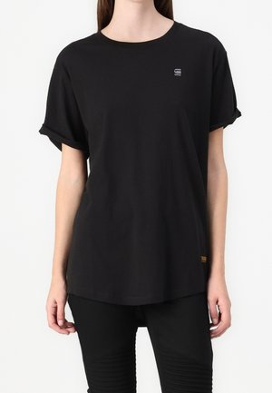 Femme portant un t-shirt noir uni à manches courtes avec un petit logo gris sur la poitrine et un pantalon noir sur fond blanc.