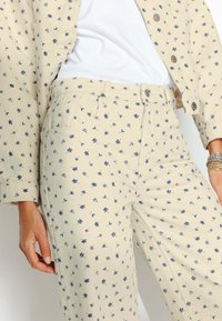 Pantalons à motif floral beige avec des motifs botaniques bleu marine, fabriqués dans un tissu doux ; dotés d'une fermeture à bouton et de poches latérales.