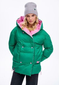 Zielona kurtka puchowa z różową podszewką, z dużymi pikowanymi sekcjami i kontrastującymi różowymi guzikami. Połączona z szarą czapką beanie.