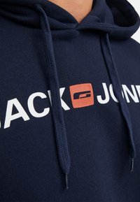 Navy blue huppari, valmistettu puuvillasta, jossa on suuri valkoinen "BLACK" ja "JON" -teksti oranssilla neliölogolla. Narut metallisine kärjineen.