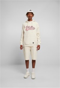 Creme hettegenser med "Phillie" grafikk i rosa, kombinert med creme shorts. Begge laget av myk tekstil. Hvite sneakers og bøttehatt fullfører stilen.