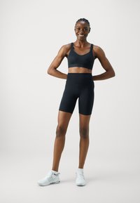 Svart sport-bh og høy midje sykkelshorts, laget av elastisk materiale, kombinert med hvite sportsko, vist mot en minimal bakgrunn.