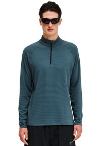 Koszula z długim rękawem, zapinana na zamek w kolorze teal, z fakturowanej tkaniny i rękawami raglanowymi. Połączona z czarnymi okularami przeciwsłonecznymi i czarnymi spodniami.