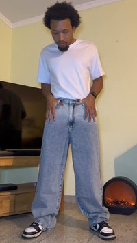 Jeans de ganga azuis claros, de perna larga, com um padrão desbotado, combinados com uma t-shirt branca de manga curta e ténis pretos. Fundo de superfície plana.