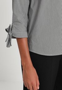 Blusa a righe bianche e nere con maniche a tre quarti e polsini annodati, caratterizzata da una trama liscia e un orlo leggermente curvo.