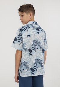 Camicia a maniche corte in tessuto azzurro chiaro con motivi floreali e foglie blu scuro, dotata di una fascia posteriore e aperture laterali per facilitare i movimenti.