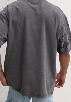 Szara, oversize koszulka z teksturowanej tkaniny. Krótkie rękawy mają czarny akcent w wzór. Rąbek jest zaokrąglony z surowymi krawędziami.