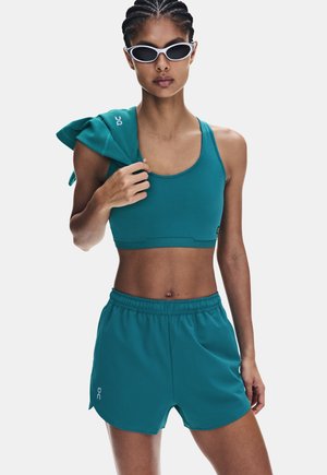 Donna atletica che indossa un reggiseno sportivo turchese e pantaloncini, tiene una giacca abbinata sulla spalla e indossa occhiali da sole con montatura bianca.