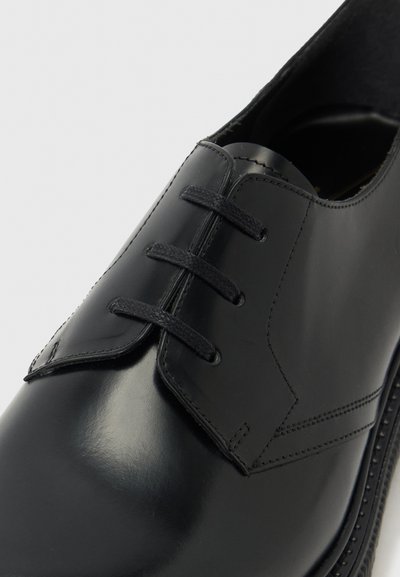 Chaussure habillée en cuir noir poli avec laçage fermé, trois œillets, et coutures détaillées sur un fond gris clair.