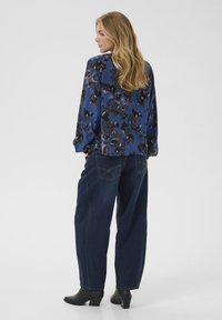 Donna con lunghi capelli biondi, che indossa una blusa floreale blu, jeans scuri larghi e stivaletti neri con tacco, di spalle.