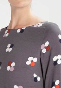 Haut gris avec un col bateau et un motif floral aux formes multicolores, comprenant des accents blancs, rouges, roses et bleu marine.