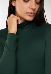 Ciemnozielony golf o dopasowanym kroju, wykonany z miękkiego, elastycznego materiału. Posiada długie rękawy i wysoki kołnierz.