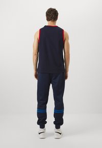 Temno moderni top brez rokavov z rdečimi odprtinami za roke, v kombinaciji z modrimi trenirkami, ki imajo modre proge blizu robov. Material se zdi mehak in udoben.