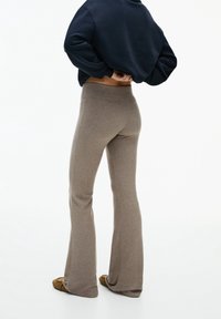 Persoon in taupe flared gebreide broek, bruine schoenen en een zwart langemouwen top, staande met de handen achter de rug.