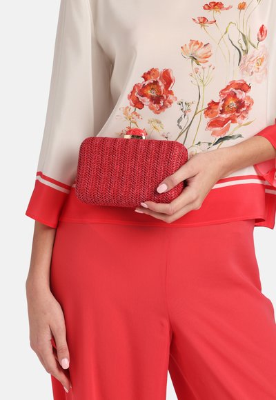 Rød clutchtaske med et tekstureret vævet design og en guldtonet spænde, holdt i hånden mod en blomsterbluse og ensfarvede røde bukser.