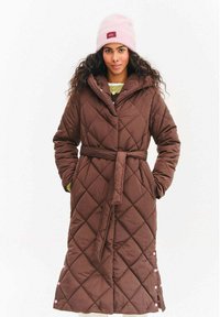 Žena v dlhom hnedom prešívanom kabáte s opaskom a vreckami, v kombinácii s ružovou čiapkou a svetlom žltým svetrom pod ním.