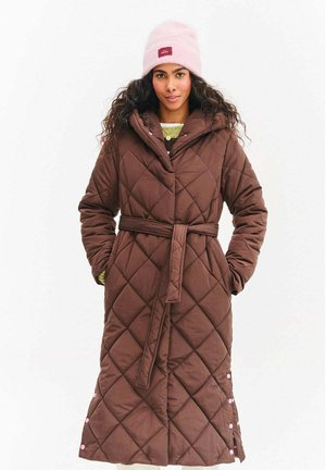 Kobieta w długim brązowym pikowanym płaszczu z paskiem i kieszeniami, w komplecie z różową czapką beanie i jasnżółtym swetrem pod spodem.