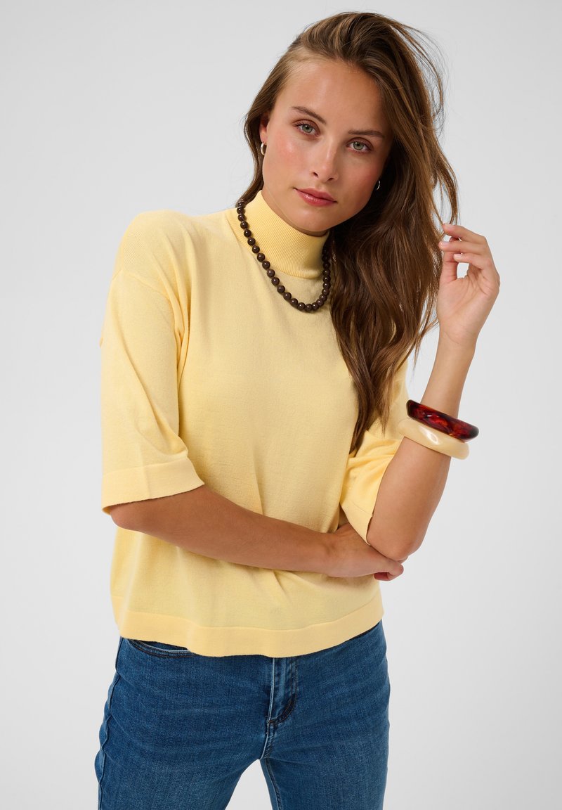 Femme aux longs cheveux bruns portant un pull à col roulé jaune à manches courtes, un jean bleu, un collier de perles marron et deux bracelets à son poignet droit.