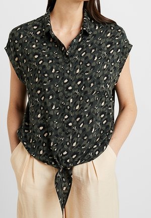 Blusa de botón sin mangas de estampado de leopardo en verde oscuro, atada a la cintura, combinada con pantalones de tiro alto en beige claro, manos metidas en los bolsillos.