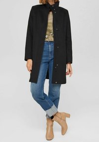 Manteau noir ajusté avec un col haut et une fermeture à boutons, associé à un jean bleu clair retroussé et des bottines beige à cheville.