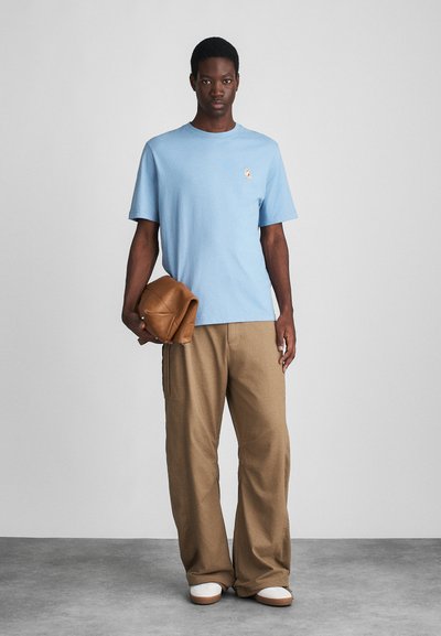 Ανοιχτό μπλε βαμβακερό t-shirt με μικρό λογότυπο στο αριστερό στήθος, συνδυασμένο με μπεζ παντελόνι φάρσα και κρατώντας μια καφέ δερμάτινη τσάντα.