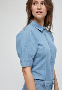 Camicia in denim azzurro chiaro con colletto, chiusura a bottoni frontale, maniche corte a palloncino e design aderente in vita. Texture morbida.