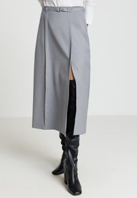 Grauer Midirock mit Frontschlitz und tailliertem Gürtel. Glatter Stoff mit strukturiertem Design. Kombiniert mit kniehohen schwarzen Lederstiefeln.