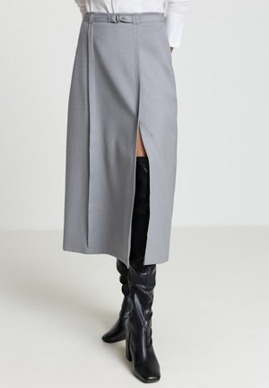 Jupe midi grise avec une fente avant et une taille ceinturée. Tissu lisse avec un design structuré. Associée à des bottes en cuir noir montantes jusqu'aux genoux.