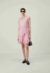 Mulher vestindo um vestido rosa com babados, óculos de sol pretos, sapatos planos pretos e segurando uma pequena bolsa preta contra um fundo simples.