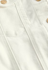 Tela de mezclilla blanca con un tejido texturizado, que presenta líneas cosidas y detalles de botones en tono dorado en la sección superior.