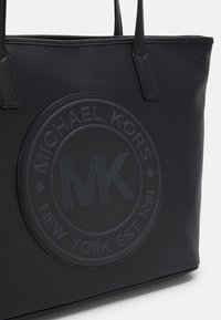 Sac en cuir noir avec un grand logo circulaire en relief portant "MICHAEL KORS" et "NEW YORK EST. 1981" dans des textures contrastées.