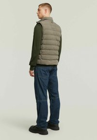 Gilet piumino grigio sopra un maglione verde, abbinato a jeans di denim blu e scarpe nere. Il gilet presenta un colletto alto e cuciture orizzontali.