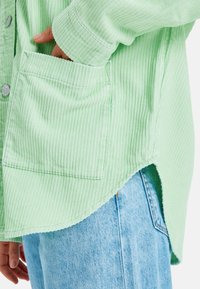 Handen i fickan på en ljusgrön ribbad skjorta som bärs över ljusblå denimjeans med böjd fåll och synliga silverknappar.