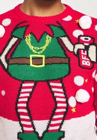 Suéter navideño rojo con piernas de elfo en medias a rayas, zapatos verdes y túnica verde con cinturón, sosteniendo una lata roja de mejor amigo con pompones blancos.