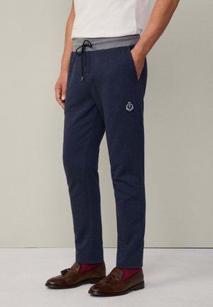 Mann in marineblauen Jogginghosen mit grauem Bund, roten Socken und braunen Tassel-Loafers, steht vor einer schlichten hellen Wand.