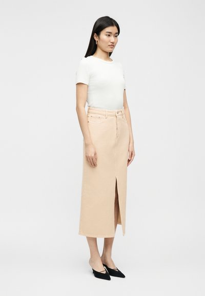 Νεαρή γυναίκα όρθια, φοράει λευκό μπλουζάκι με κοντά μανίκια, μπεζ midi φούστα με ψηλή μέση και ανοίγματα μπροστά, καθώς και μαύρες γόβες με μυτερή μύτη.