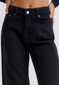 Vaqueros de cintura alta en color negro, hechos de algodón. Entre sus características se incluyen un cierre frontal con botón, dos bolsillos y detalles en metal de color cobre.
