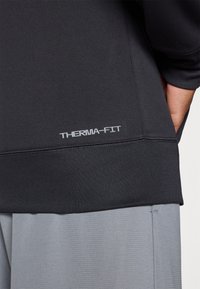 Juodas Therma-Fit megztinis su lygiu paviršiumi, su pilku logotipu ant apvado. Suderintas su šviesiai pilkomis tekstūruotomis šortais.